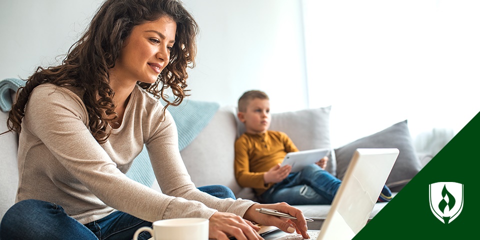 一张妈妈在用笔记本电脑做作业的照片，她坐在沙发上，旁边的儿子在用平板电脑